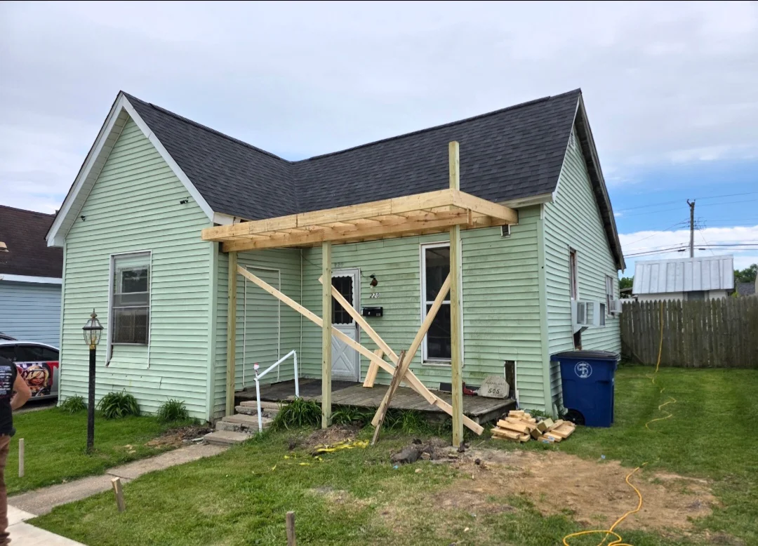 Wood pergola and covered porch with cross-bracing on green vinyl-sided house