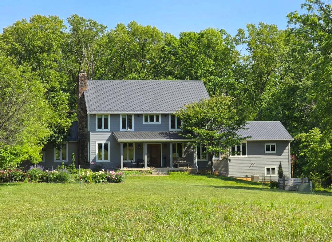 Standing seam metal roof installation on two-story farmhouse with gray metal roofing and white siding
